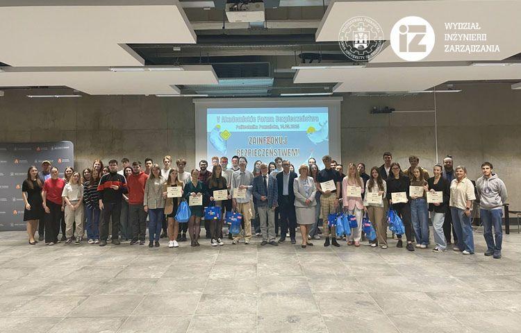 Zdjęcie grupowe uczestników 5. Akademickiego Forum Bezpieczeństwa. Licznie zgromadzeni i uśmiechnięci uczestnicy Akademickiego Forum Bezpieczeństwa stoją w szpalerze w Auli WIZ. W tle znajduje się grafika promująca wydarzenie, na której na szarym tle wyeksponowane są foki.